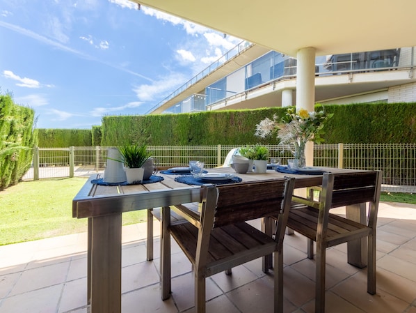 An outdoor patio with a table set for dining, surrounded by chairs, and a view of a modern building with a green hedge in the background.