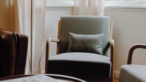 A room with a round wooden table, a grey armchair with a cushion, and a floor lamp.