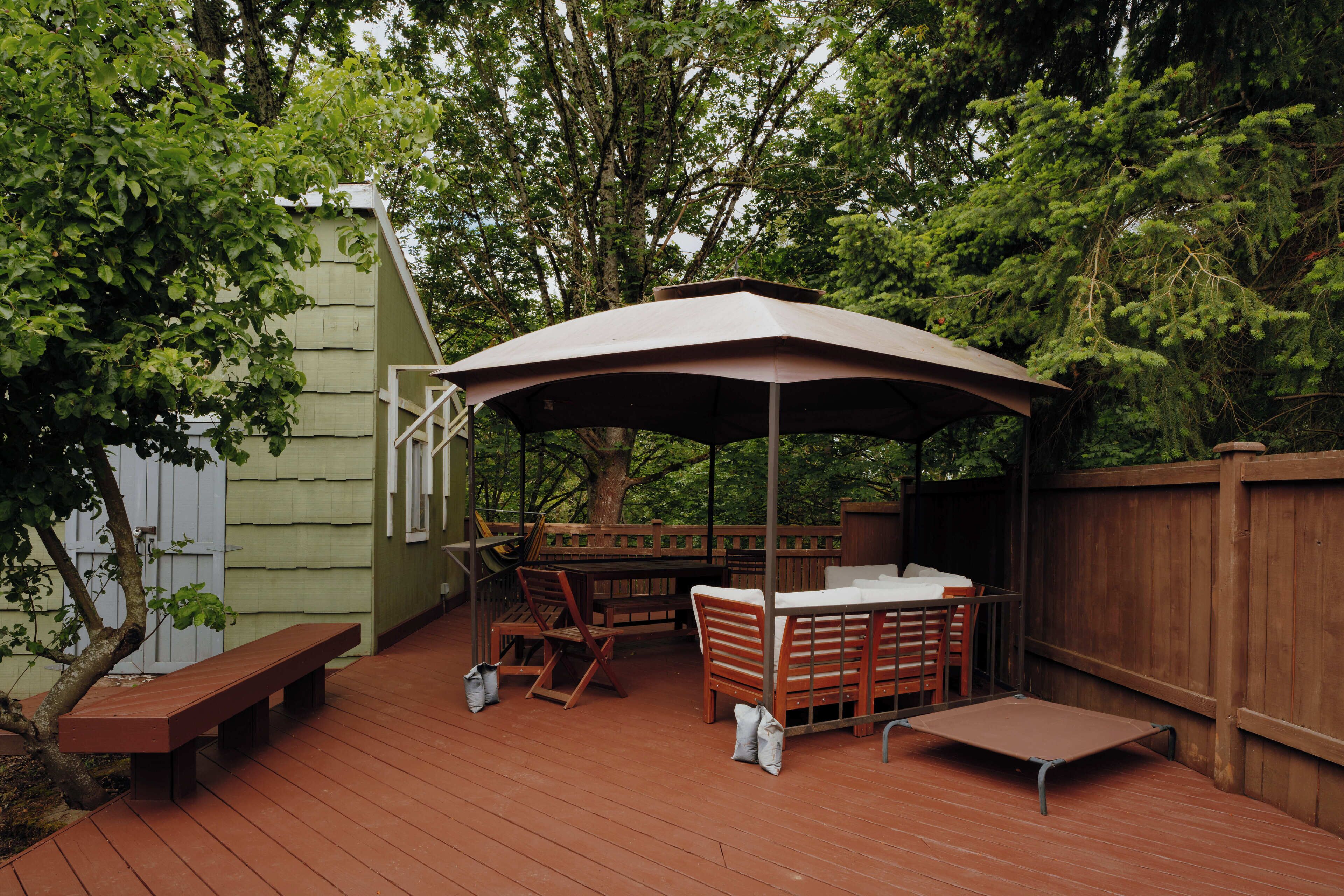 Una terraza de madera con una mesa y sillas bajo un gran paraguas, rodeada de árboles y una valla de madera.