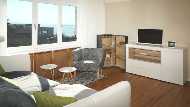 A modern living room with a sofa, armchair, and small tables. A television is on a cabinet, and there is a view of the sea through large windows. - 1 Bedroom Apartment in Lochau (Lochau)