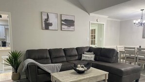 Un salon avec un canapé d’angle en cuir foncé, une table basse en bois clair sur laquelle se trouve un bol rempli d’objets décoratifs, une plante en pot et un lustre.