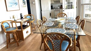 Een eetgedeelte met een glazen tafel, blauwe en witte borden met een patroon, rieten stoelen en een keuken op de achtergrond.