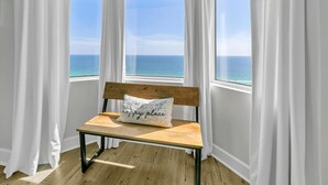 A room with a wooden bench and a pillow with text, large windows with white curtains, and a view of the ocean. - Tidewater 1802 (Panama City Beach)