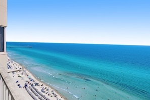 A beach with clear turquoise waters, a pier in the distance, and a balcony with sun loungers.