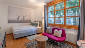 Ein modernes Schlafzimmer mit einem Bett, einem rosa Sofa, einem runden Holztisch und einem großen Fenster mit Blick auf Bäume.