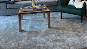 Un salon moderne avec un canapé gris, un fauteuil vert et une table basse. Un tableau représentant un coucher de soleil est accroché au mur, et la pièce est dotée d’un plafond en bois.