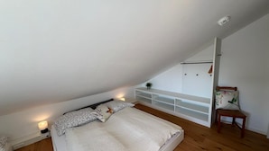 Une chambre avec un plafond en pente, un lit avec une literie à motifs, une table de chevet avec une lampe, une petite chaise en bois et un meuble de rangement blanc adossé au mur.