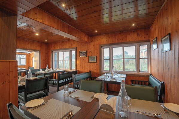 A wooden-paneled dining area with tables set for two, benches, and large windows offering a view of the outdoors.