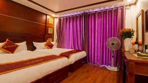 A hotel room with two beds, wooden headboard, purple curtains, a bedside table with a fan, and a vase with flowers.