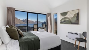 Une chambre d’hôtel avec un grand lit, une vue sur les montagnes et un lac, un tableau au mur et un fauteuil.