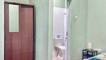 A bedroom with a bed, a wardrobe, a bathroom with a toilet and sink, and a small table with a lamp.