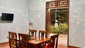 A dining area with a wooden table and chairs, a TV mounted on the wall, and a view of greenery through a window. - Telu Telu Solo (Surakarta)