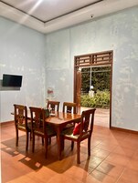 Uma área de refeições com mesa e cadeiras de madeira, uma TV fixada na parede e vista para o verde através de uma janela.