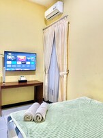 Quarto de hotel com uma cama, televisão sobre um suporte de madeira, ar condicionado e cortina.