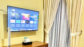 A flat-screen TV mounted on a wooden desk with a remote control, a tissue box, and a water bottle on it. The room has a yellow wall and a window with beige curtains. - Telu Telu Solo (Surakarta)