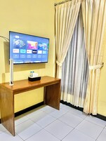Uma TV de tela plana fixada em uma escrivaninha de madeira, com um controle remoto, uma caixa de lenços de papel e uma garrafa de água sobre ela. O quarto tem uma parede amarela e uma janela com cortinas bege.