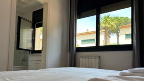 A hotel room with a bed, a window with a view of buildings and palm trees, a radiator, and a bathroom with a shower. - Preciosa Casa, con Enorme Piscina en Zona Tranquila y Familiar (Sant Feliu de Guixols)