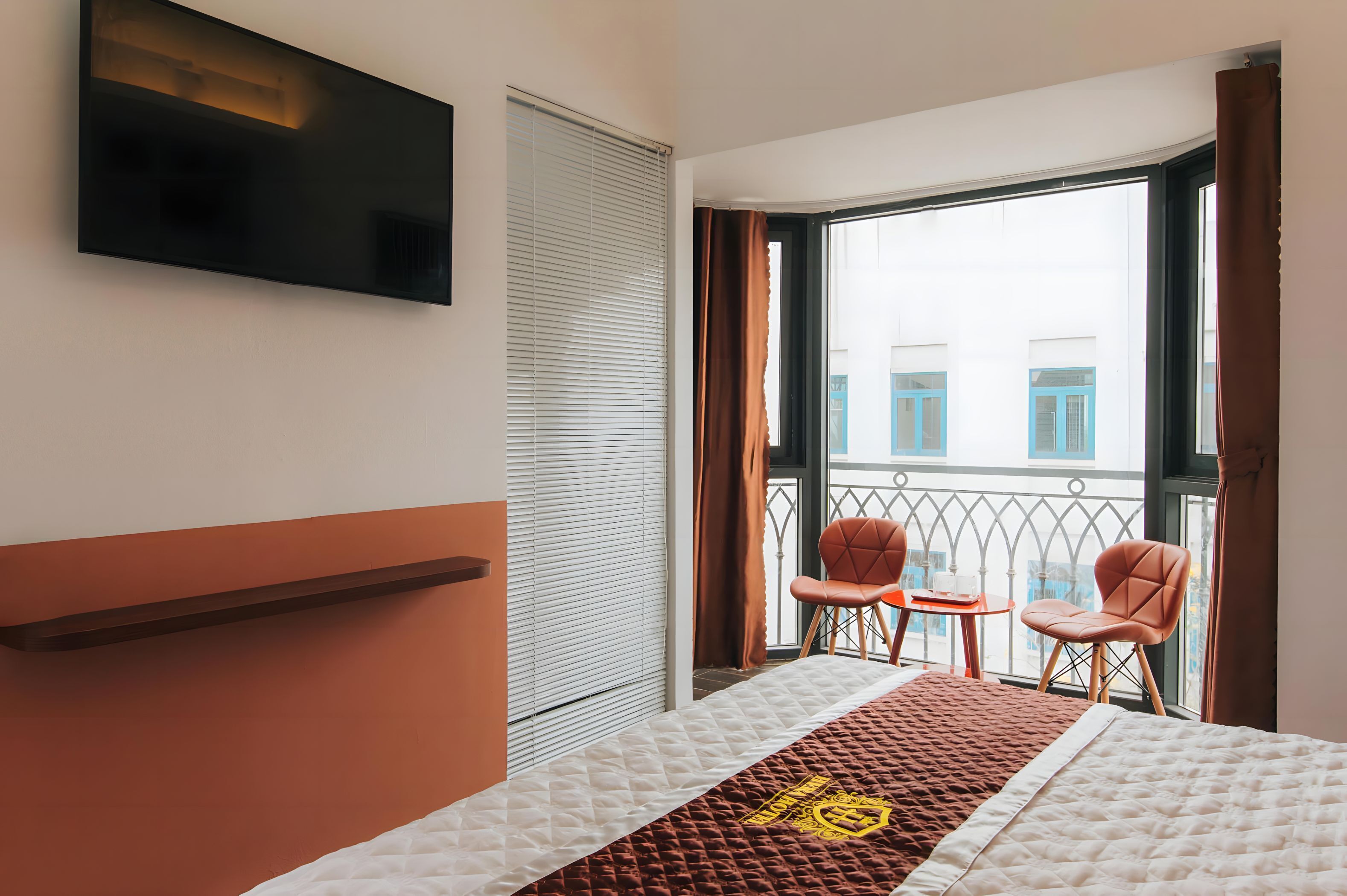 A hotel room with a bed, a TV mounted on the wall, a balcony with two chairs, and a view of buildings outside.