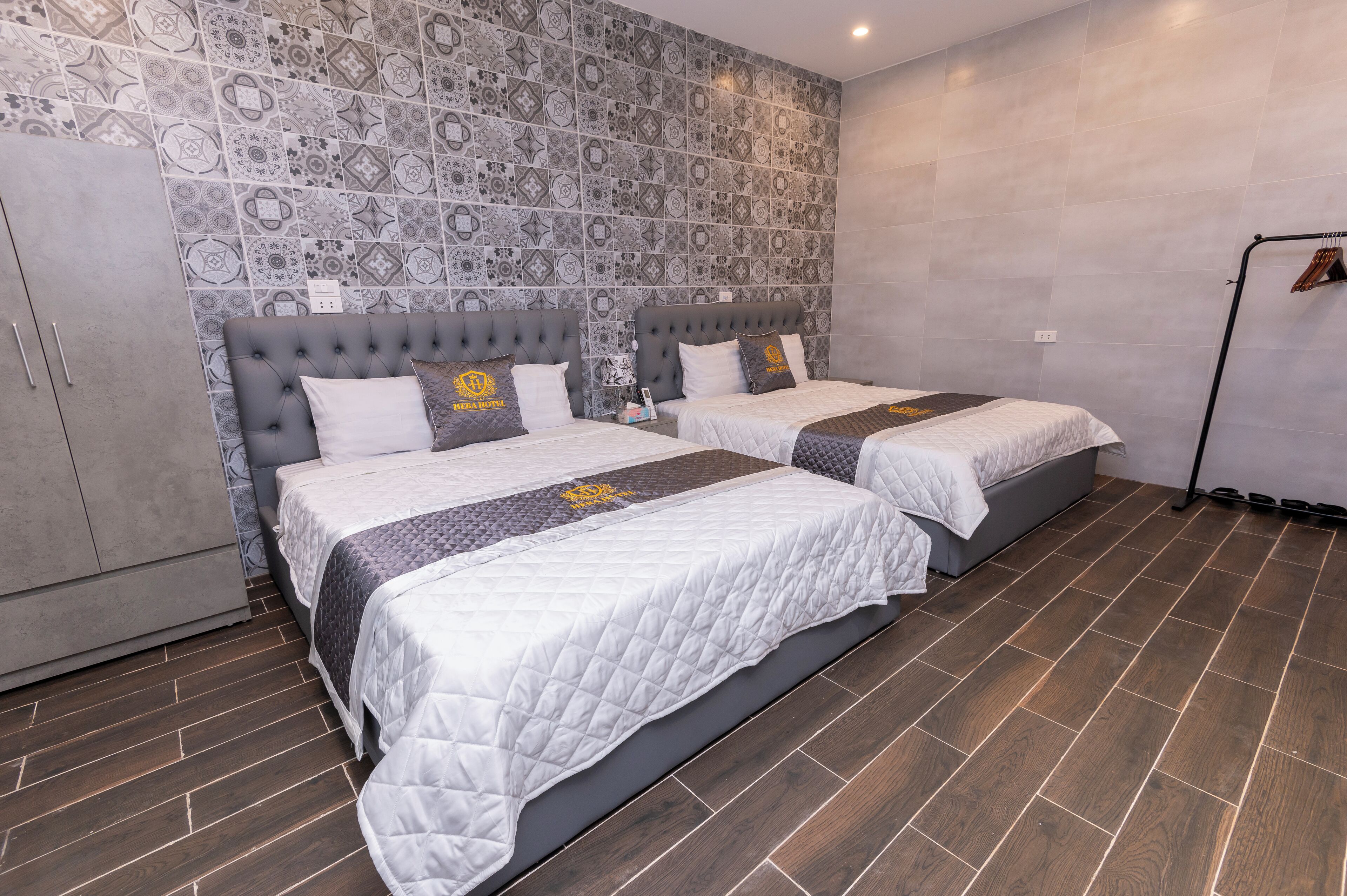 A hotel room with two beds, grey headboards, patterned wallpaper, and a grey wardrobe.