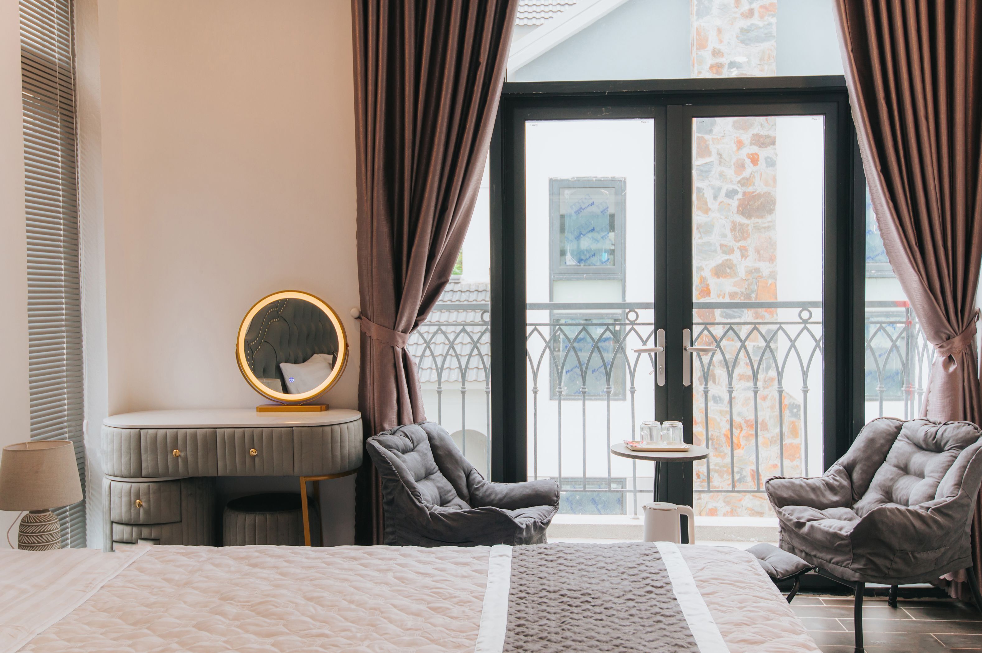 A bedroom with a bed, two armchairs, a vanity with a mirror, and a balcony with a railing.