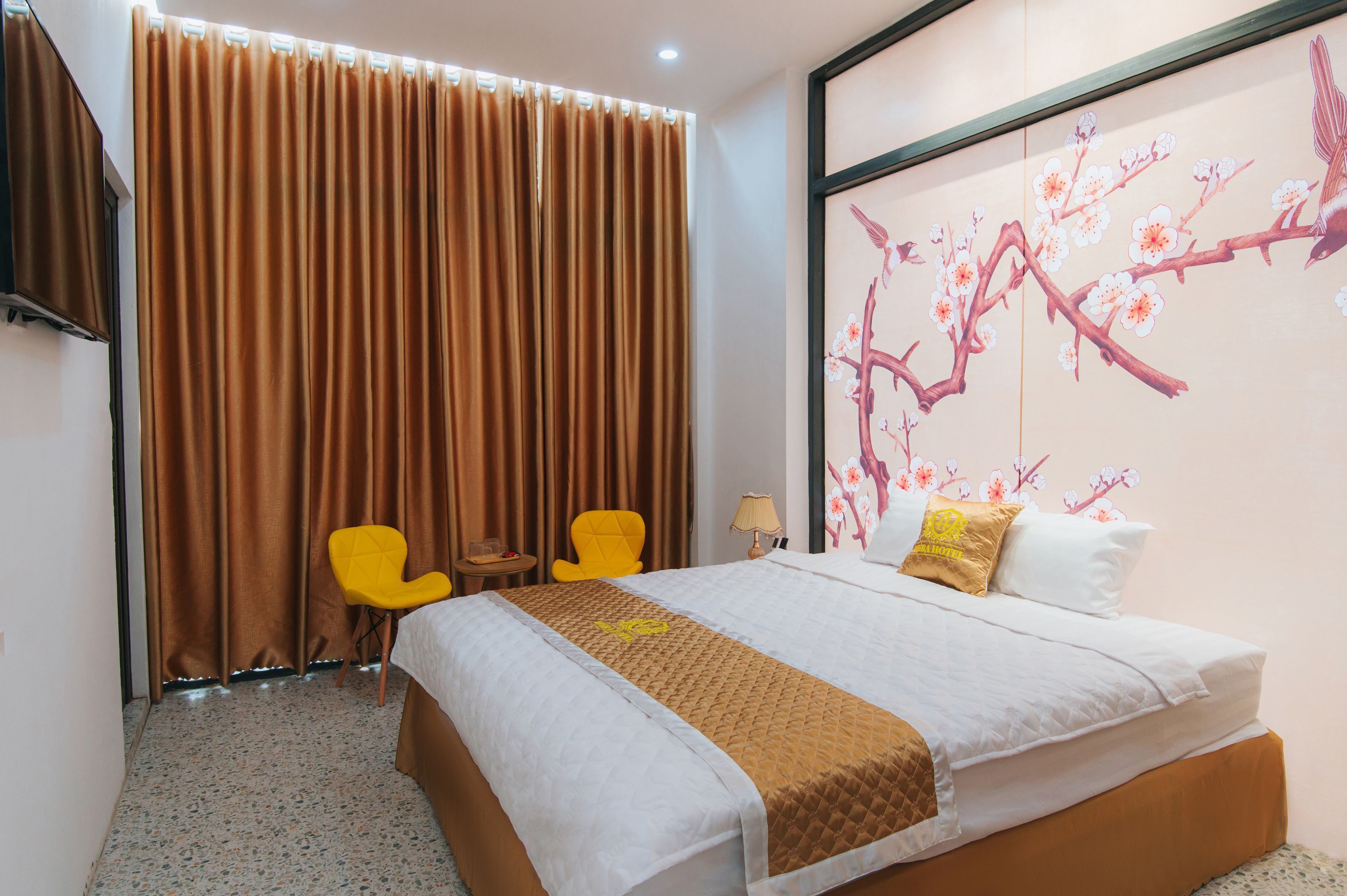 A hotel room with a bed, curtains, a yellow chair, a small table, and a wall with a floral painting.