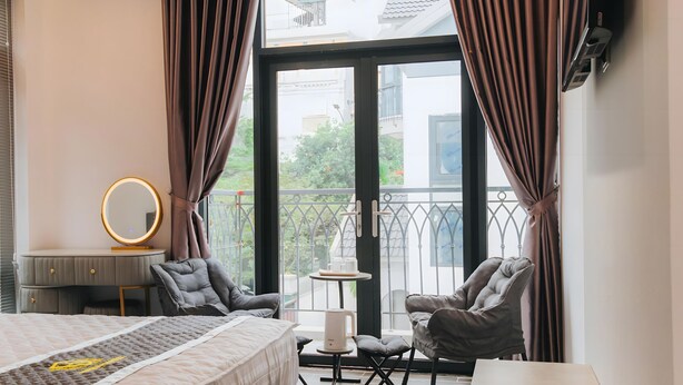 Una camera d'albergo con un letto, una toeletta con uno specchio, due poltrone, un piccolo tavolino con una lampada e un balcone con vista sull'esterno.