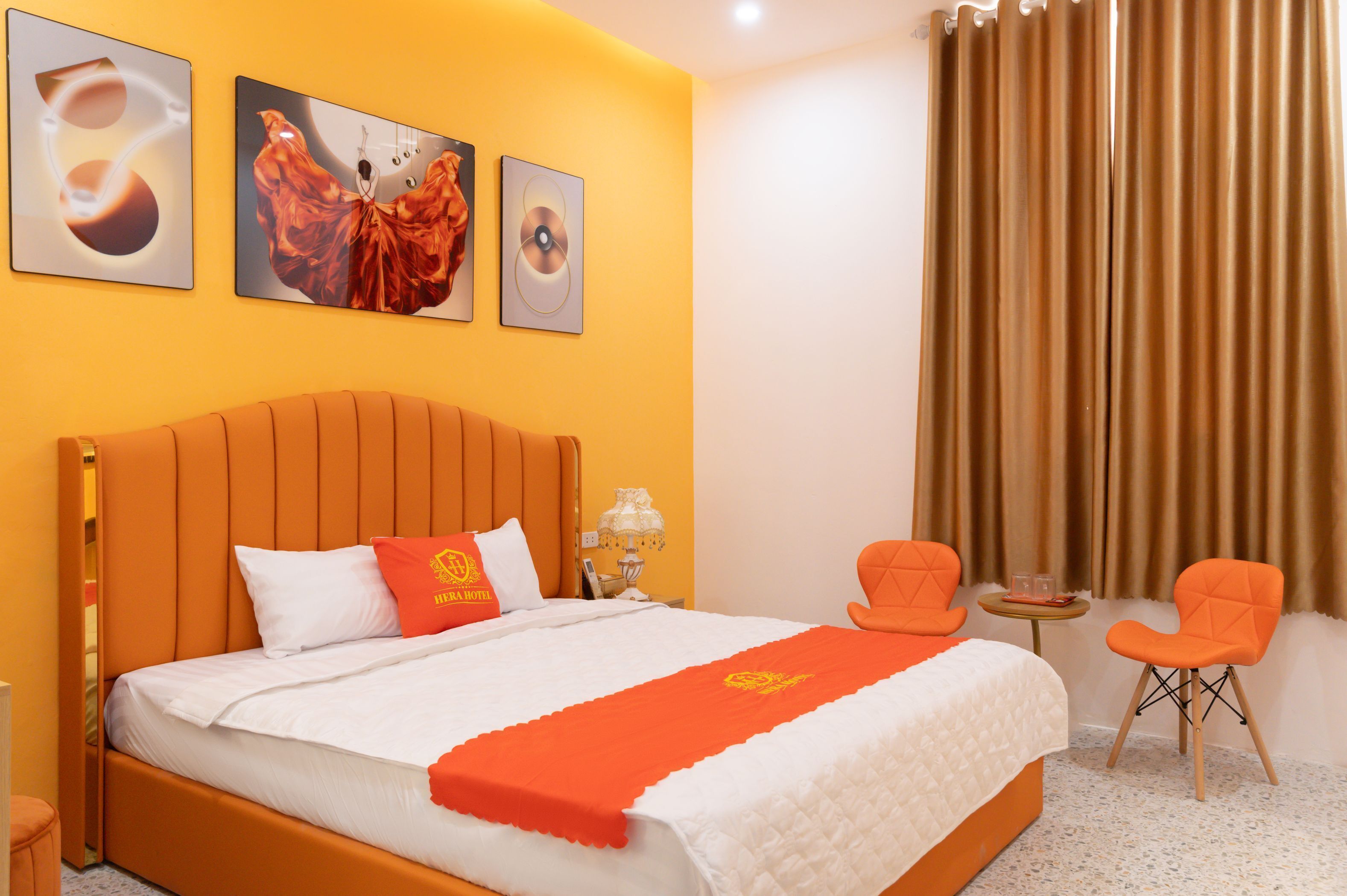A hotel room with a large bed, orange headboard, and a red throw blanket. There are two orange chairs and a small table with a lamp.