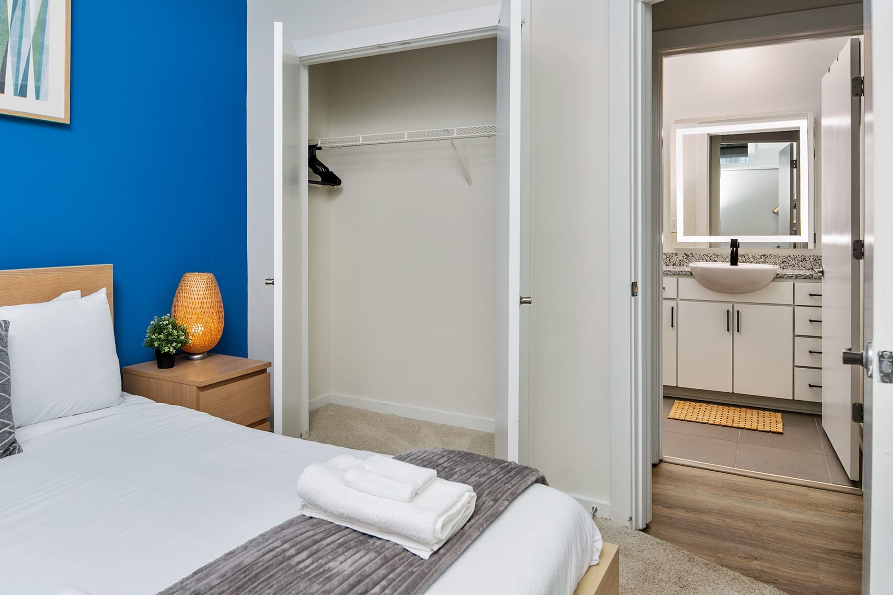Habitación de hotel con cama, mesita de noche, lámpara, armario y baño con lavabo y espejo.