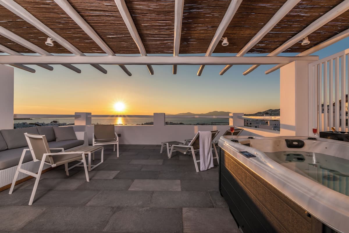 A rooftop terrace with a sofa, chairs, a hot tub, and a view of the sunset over the sea.