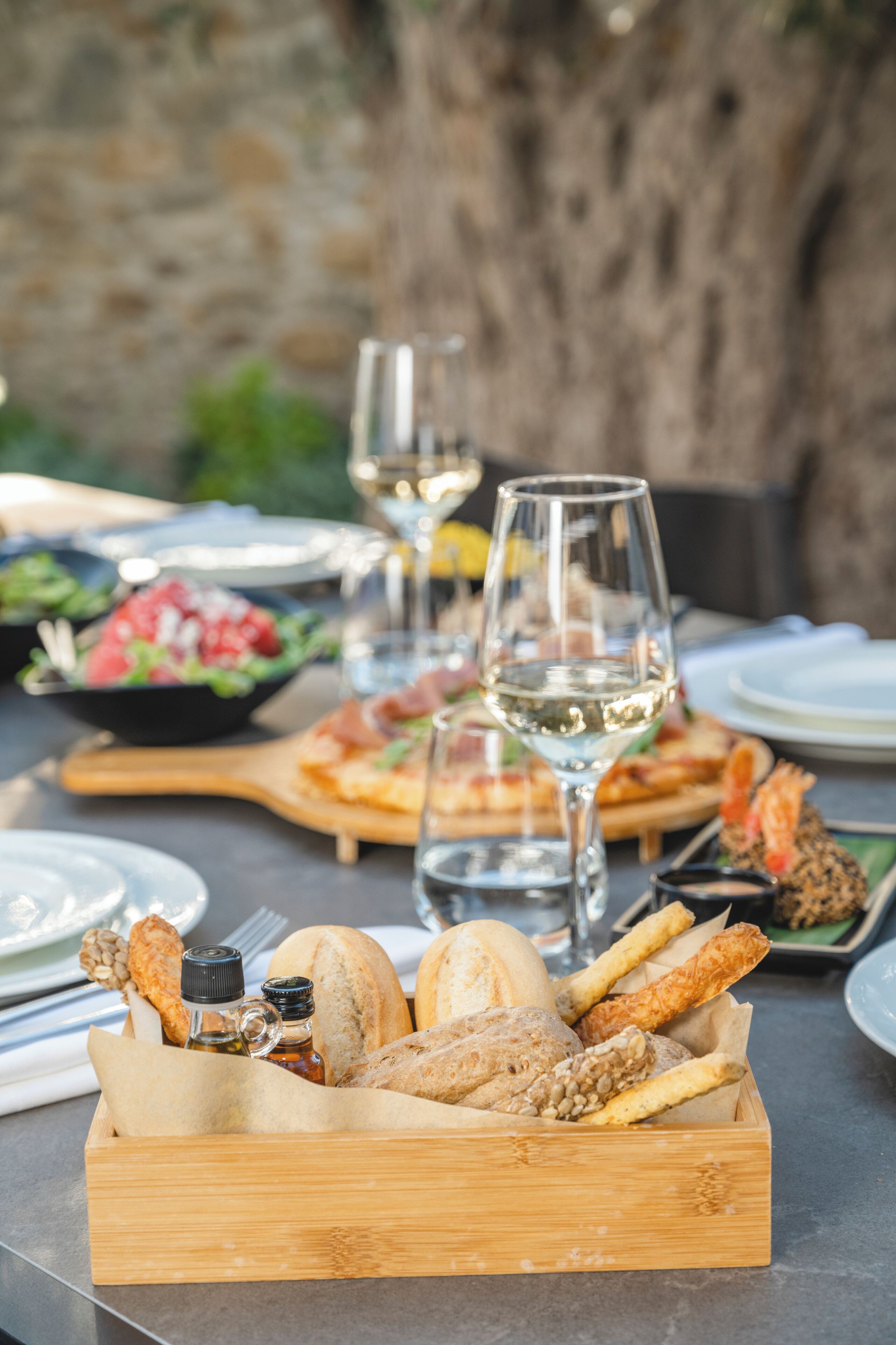 A wooden box filled with various types of bread and crackers, accompanied by a glass of white wine on a table set for a meal.