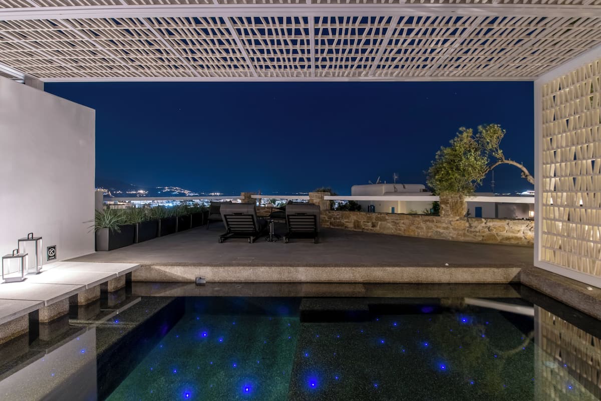 An outdoor pool area with a view of the city at night, a pergola, and a seating area with chairs and a table.