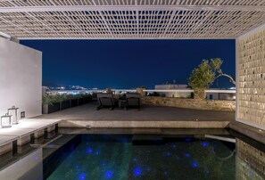 An outdoor pool area with a view of the city at night, a pergola, and a seating area with chairs and a table. - Rochari  (Mykonos)