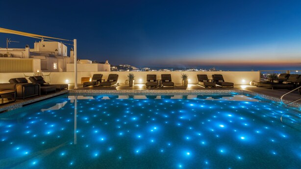 Piscina en la azotea con luces LED azules, sillones de descanso y vistas a la ciudad al atardecer.