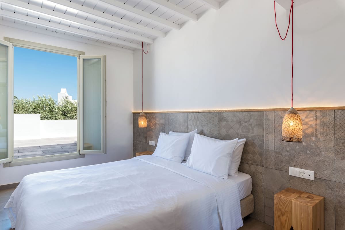 A bedroom with a large bed, a wooden nightstand, a wall-mounted light fixture, and a view through an open window.