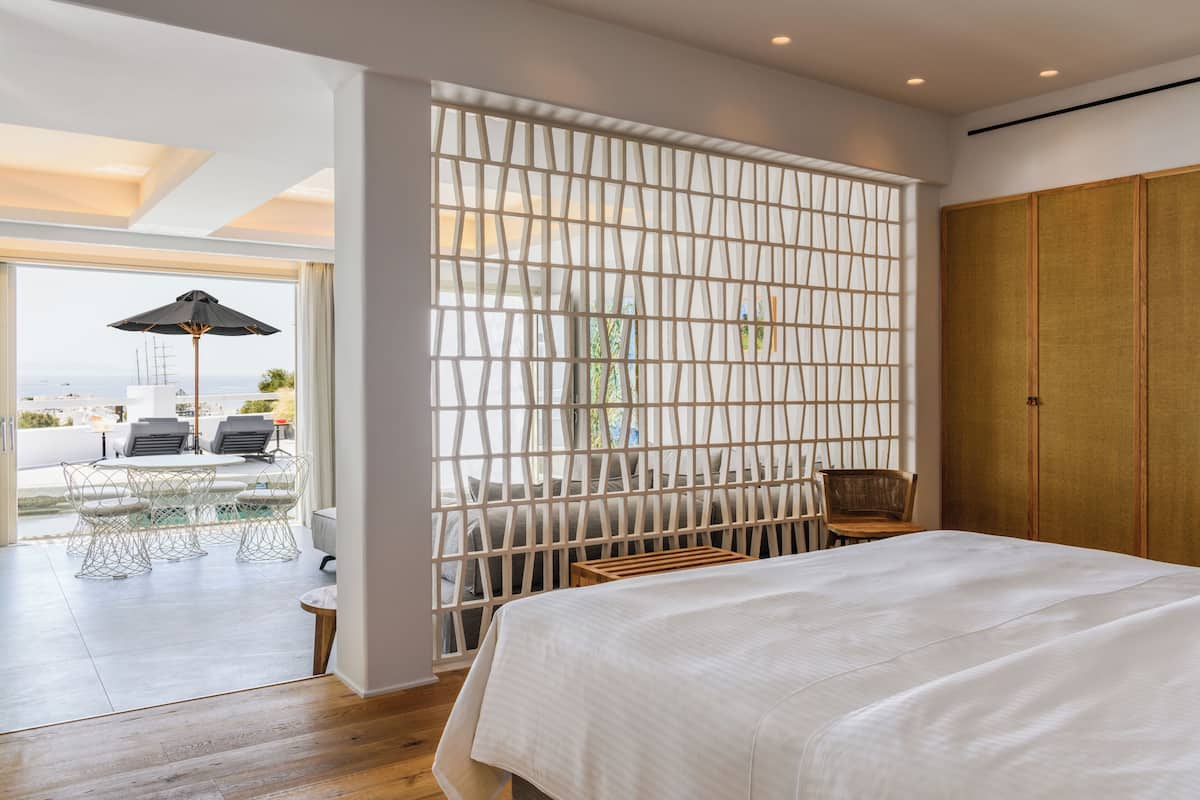 A modern hotel room with a large bed, a view of the outdoors, a balcony with a table and chairs, and a wooden wardrobe.