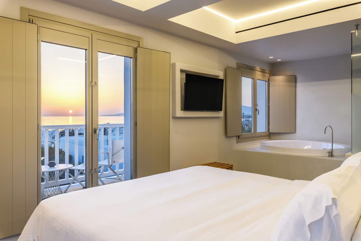 A modern hotel room with a large bed, a TV mounted on the wall, a bathtub, and a balcony with a view of the sunset.