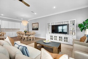 Un salon moderne avec un coin repas, des meubles blancs, un grand téléviseur et un lustre.