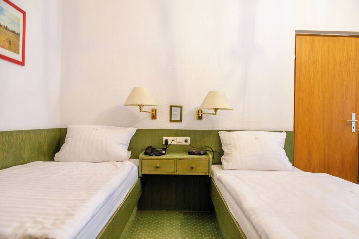 Une chambre d’hôtel avec deux lits simples, une tête de lit verte, des lampes de chevet, une table de chevet avec un téléphone et une télécommande, et une porte en bois.