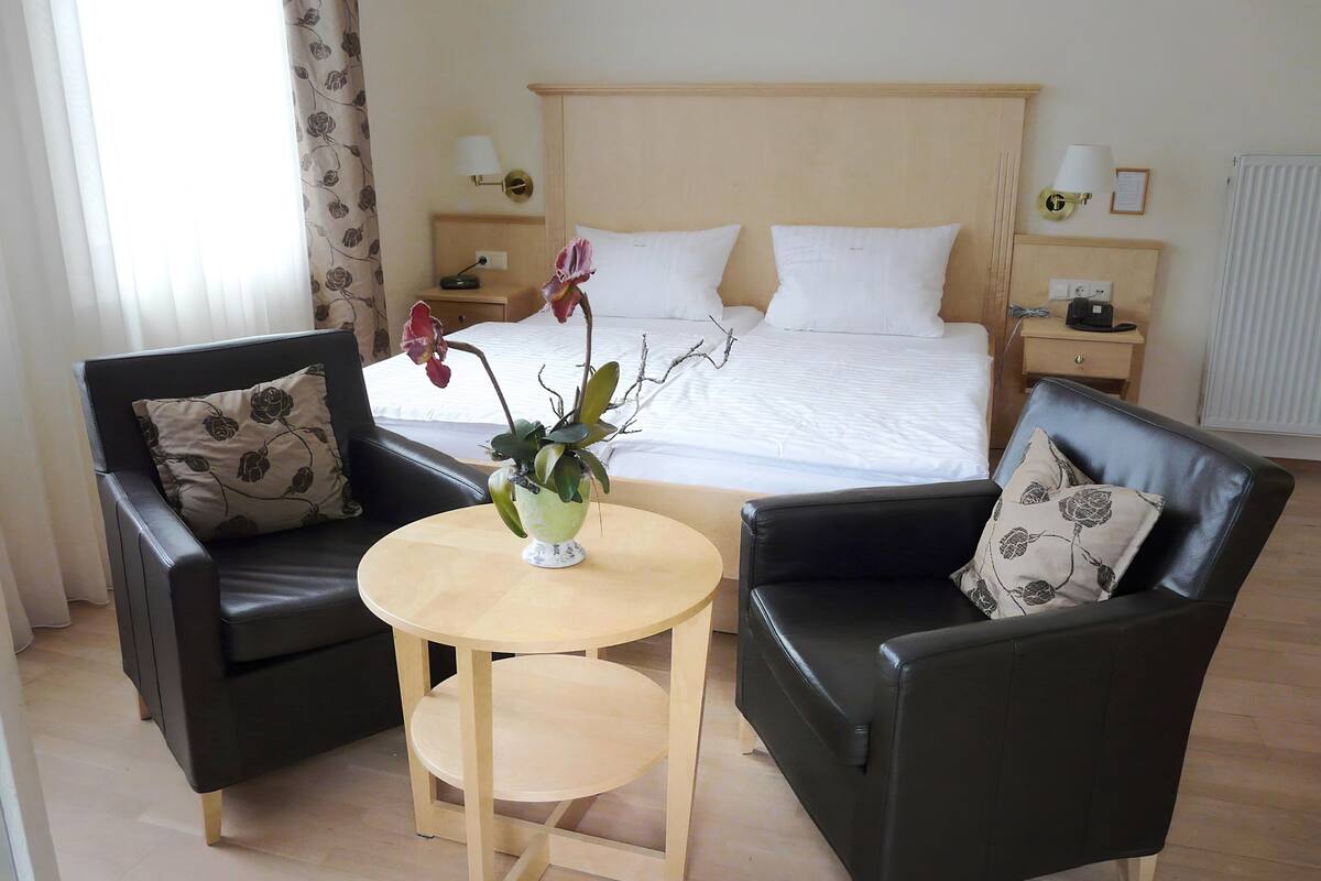 Une chambre d’hôtel avec un lit, deux fauteuils en cuir noir, une petite table en bois ornée d’un vase de fleurs et une table de chevet avec une lampe et un téléphone.