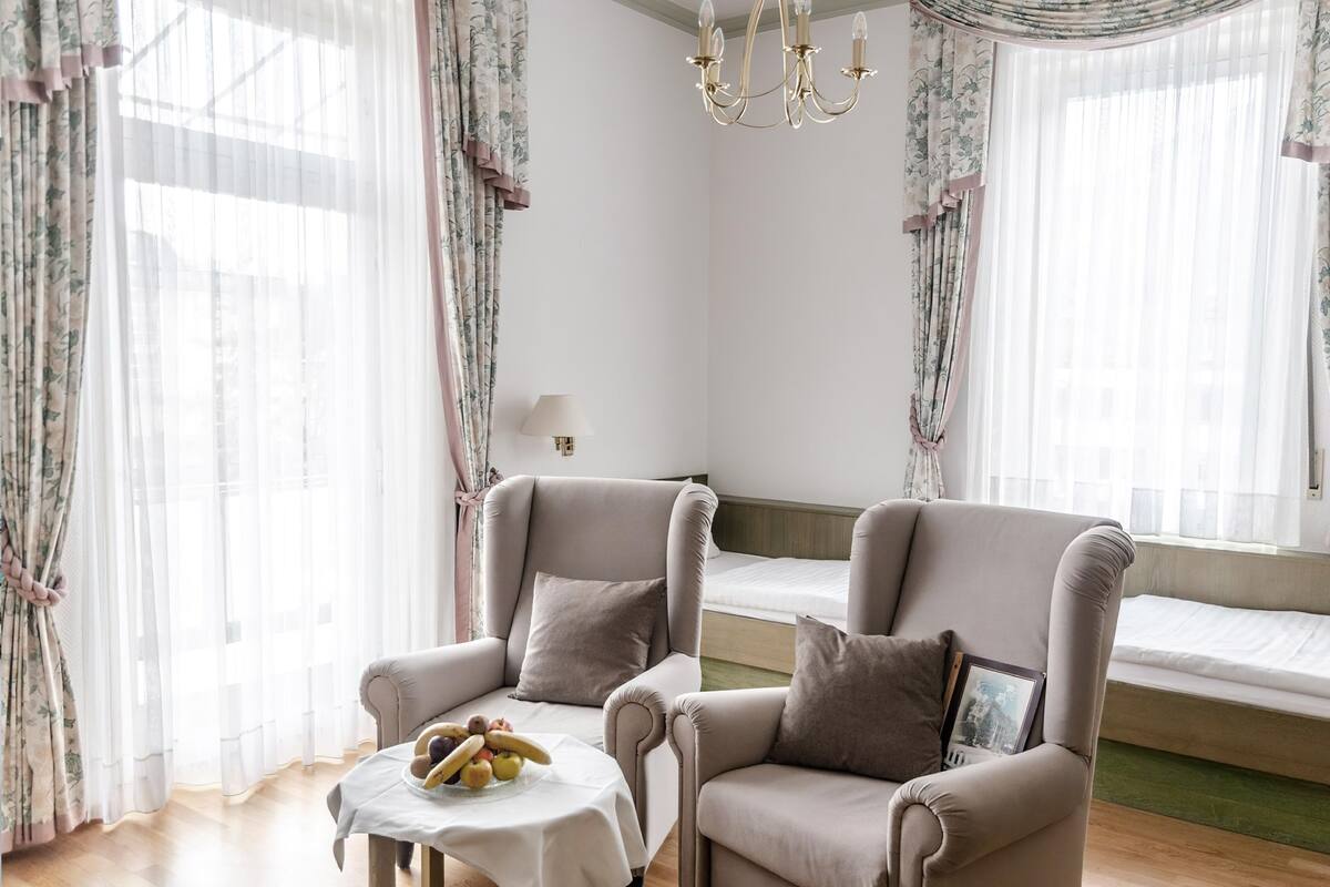 Une pièce comprenant deux fauteuils, une petite table avec des fruits, un lit recouvert d’une couverture verte et un lustre.