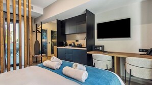 A modern hotel room with a bed, a desk, a chair, a TV, and a kitchen area. - Mc Queen Apartments (Athens)