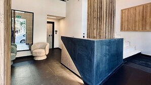 A modern reception area with a dark reception desk, a wooden accent wall, and a staircase leading to an upper level. - Mc Queen Apartments (Athens)