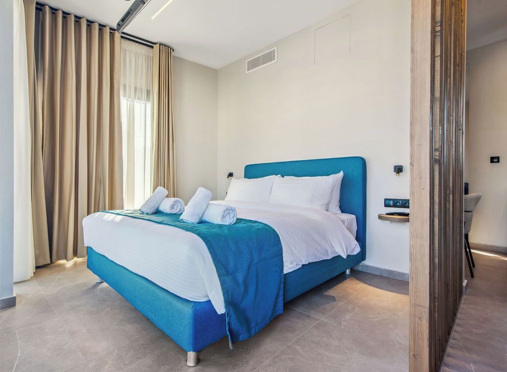 A modern hotel room with a blue bed, white bedding, and beige curtains.