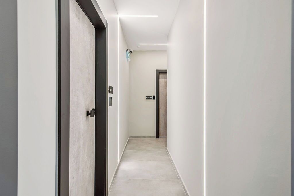 Un pasillo moderno con piso y paredes de tonos claros, una puerta a la izquierda y un ascensor a lo lejos.