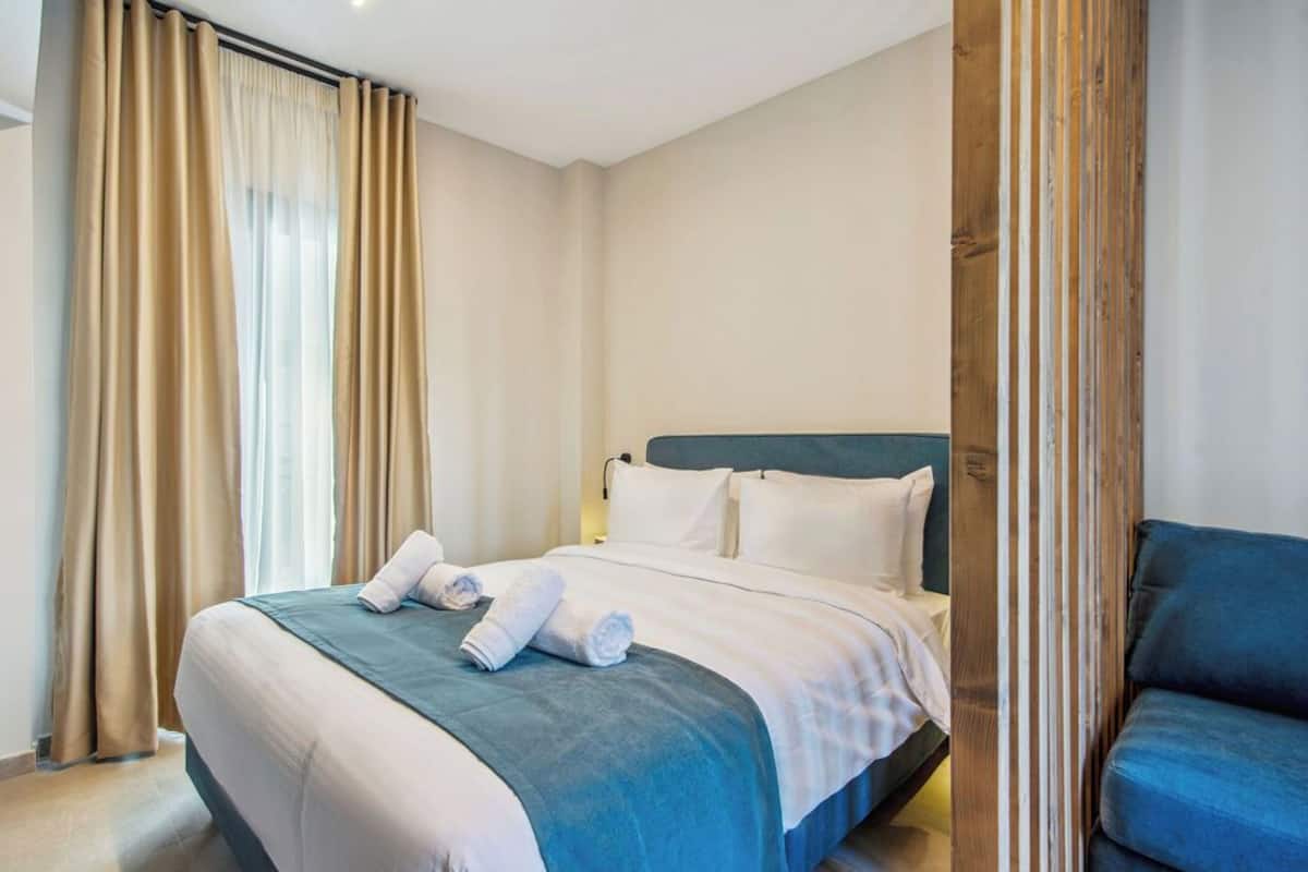 Een netjes opgemaakt bed met een blauwe plaid en witte kussens, een houten hoofdbord, en een blauwe fauteuil aan de rechterkant.