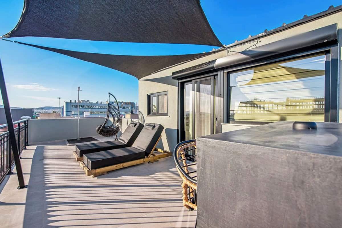 Een zonnig terras buiten met lounge stoelen, een tafel en een groot raam met uitzicht op een stadsbeeld.