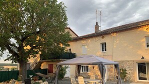 A covered outdoor seating area with a table and chairs, a stone wall, and a large tree. - Chez mamie (Montdragon)