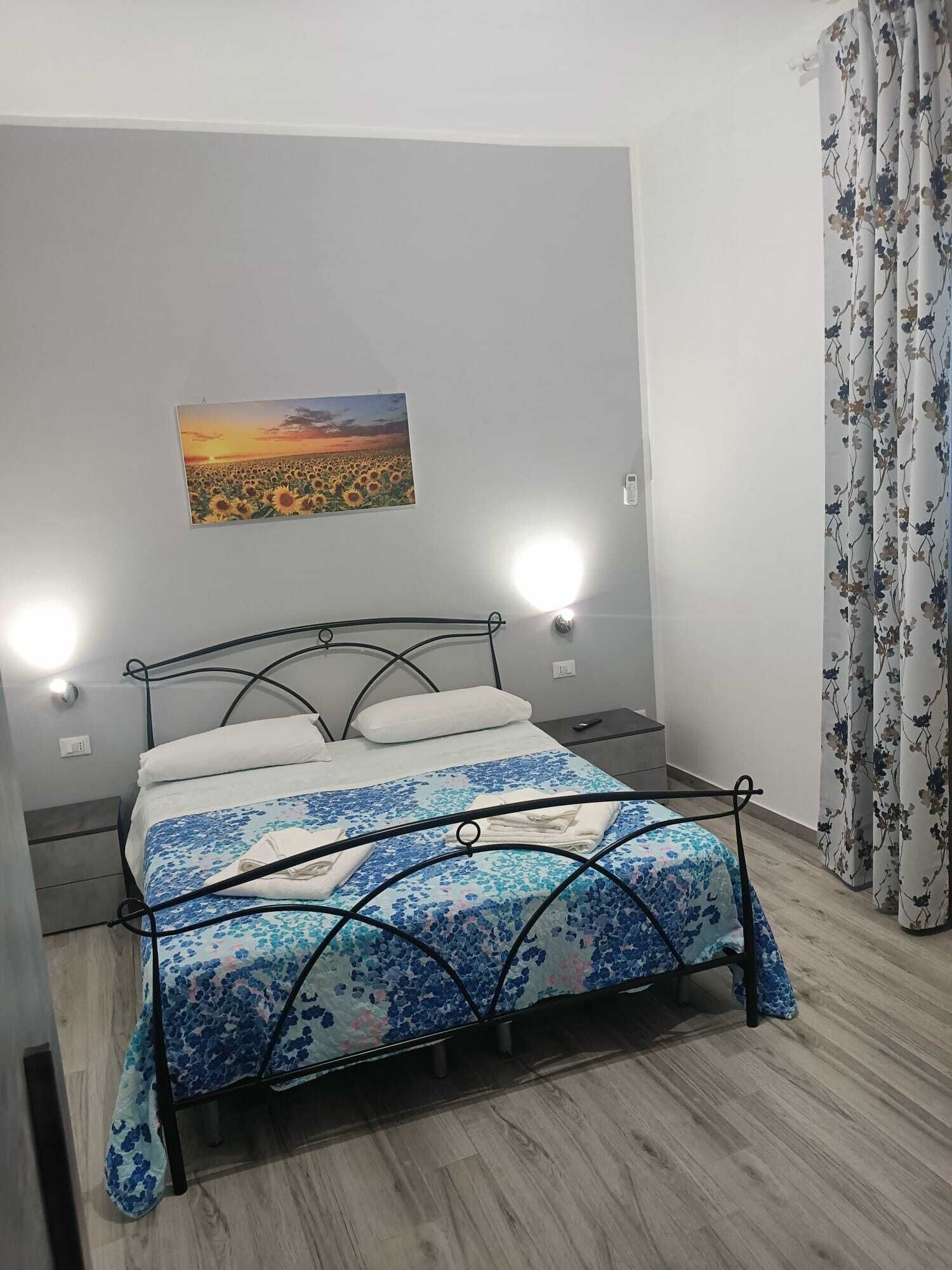 A bedroom with a bed, a nightstand, a wall-mounted light, a painting, and a curtain.