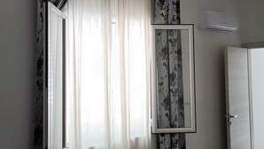 Une pièce dotée d’une grande fenêtre ornée de rideaux blancs et vaporeux et de tentures à motifs floraux, d’un tableau encadré accroché au mur et d’une porte située à droite.