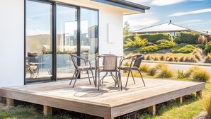 Una casa moderna con terraza de madera, dos sillas de mimbre y una puerta de cristal que da a un jardín.